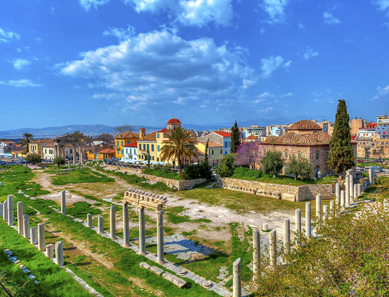 Ancient stone ruins with columns in a grassy field, surrounded by colorful town buildings and a domed structure under bright blue sky