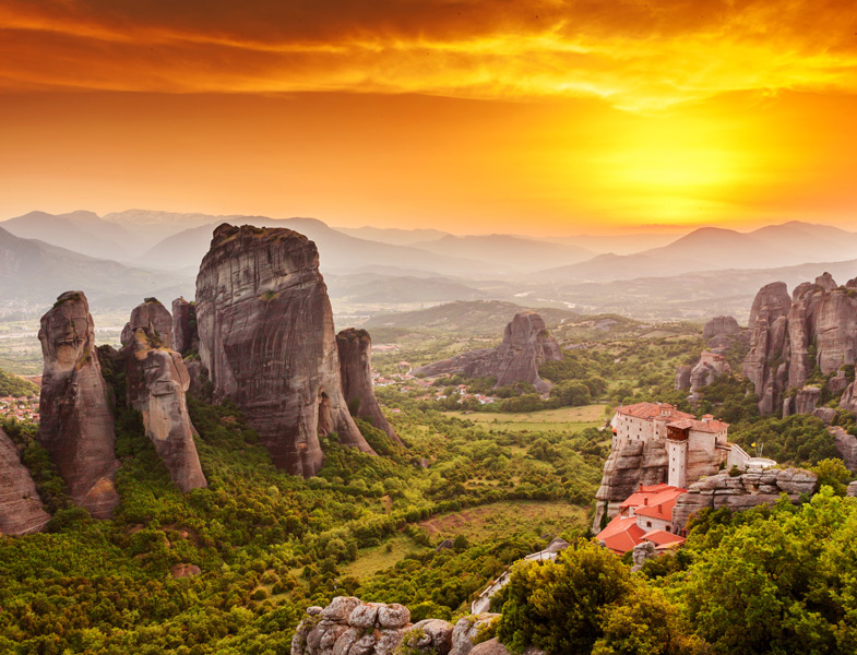 Dramatic sunset over tall rock formations with a monastery building perched on a cliff, surrounded by green valley and mountains
