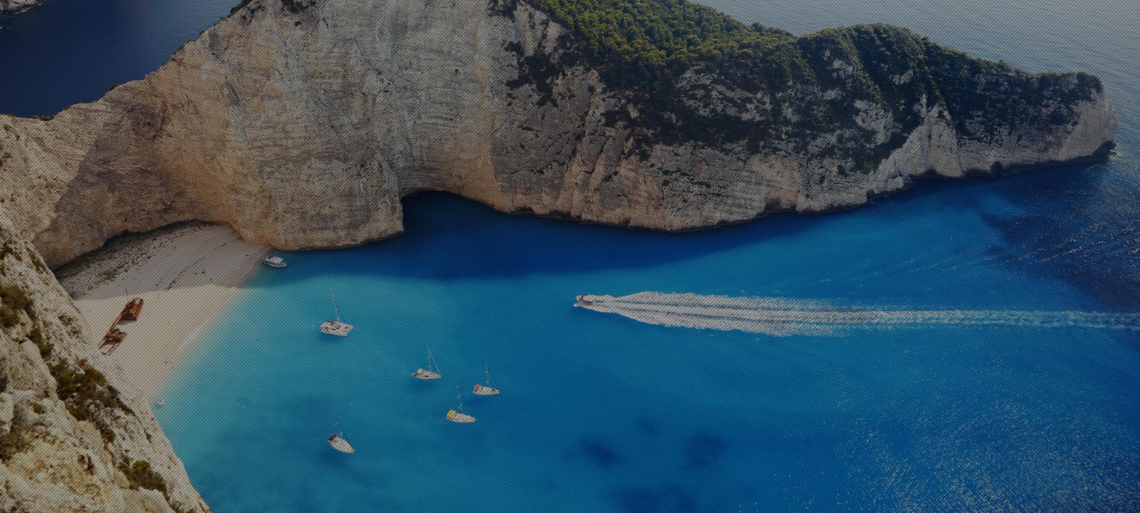 Aerial view of a turquoise bay with steep cliffs, a sandy cove, sailboats, and a speedboat leaving a wake