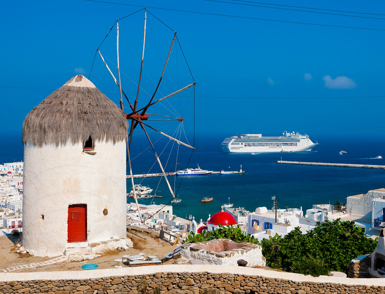 Traditional white windmill with thatched roof on a hillside overlooking a blue harbor with boats and a large cruise ship