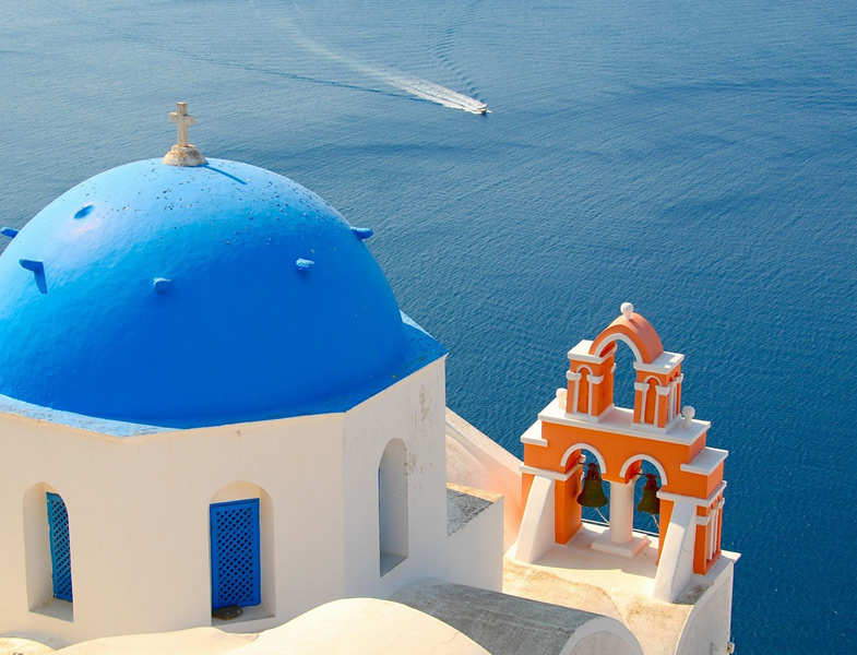 Close-up of a white building with a blue domed roof and an orange bell tower overlooking deep blue sea, a small boat passing by