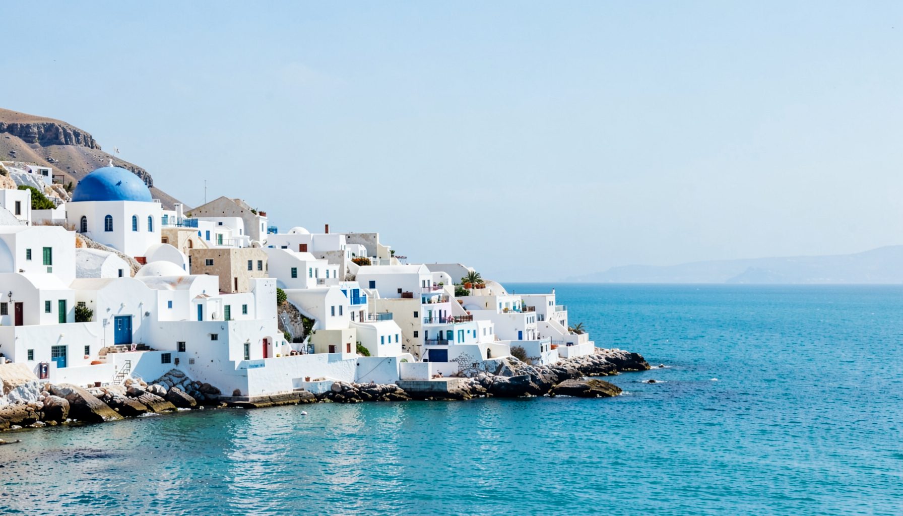 Sunlit whitewashed coastal village with blue-domed buildings overlooking a calm turquoise sea